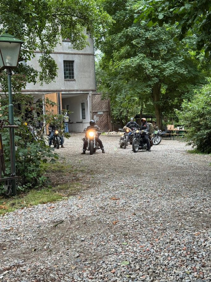 macht euch mit eurem Naturwohnzimmer auf den Weg Zwei Motorräder stehen auf einem Kiesweg neben einem älteren Gebäude und Bäumen.