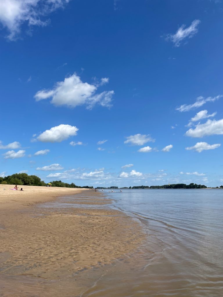 Heller Sandstrand unter blauem Himmel mit vereinzelten Wolken und ruhigem Wasser.