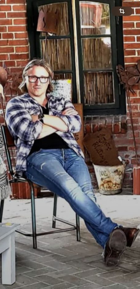 The Man Person mit Brille und kariertem Hemd sitzt entspannt auf einem Stuhl.